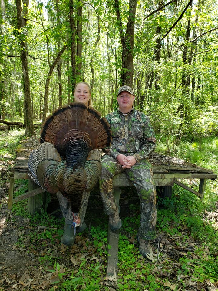 Scott Phillips and woman, in hunting gear post in the woods with a harvested wild turkey.