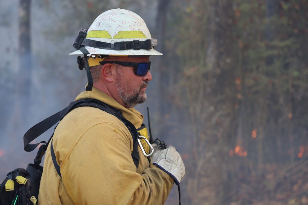 wildland firefighter wearing PPE, smoke and flame in the background