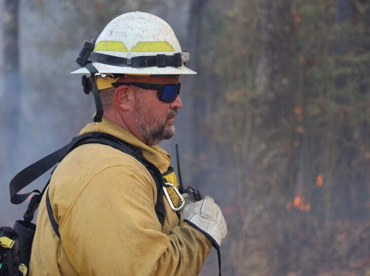 wildland firefighter wearing PPE, smoke and flame in the background