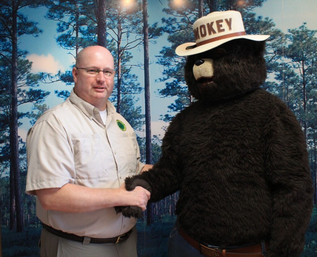 Scott Phillips, SC State Forester shakes hands with Smokey Bear mascot in front of printed forest backdrop 