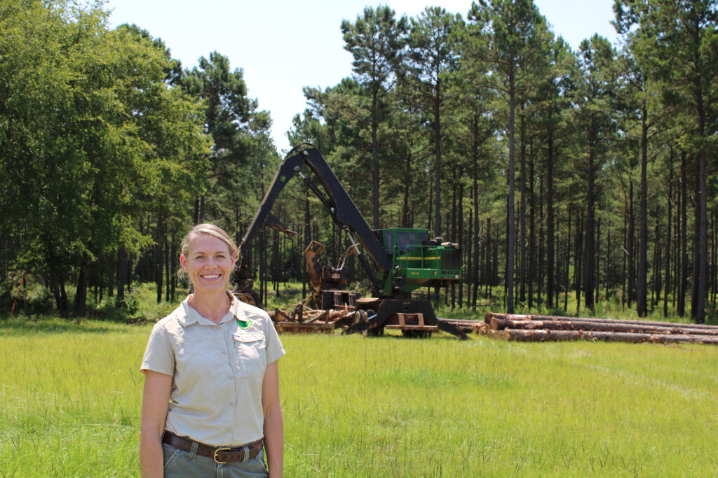 Female forester stands smiling in foreground, with a logging operation occurring in the woods behind her