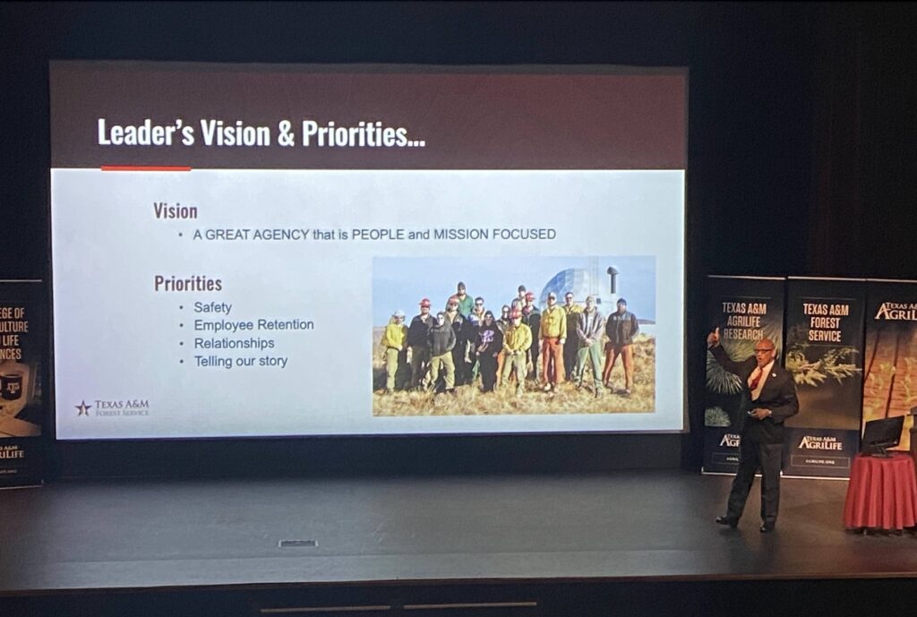 Al Davis giving a presentation on a stage, him gesturing to a projected image of a PowerPoint slide that says "Vision: A great agency that is people and mission focused. Priorities: Safety, Employee Retention, Relationships, Telling Our Story