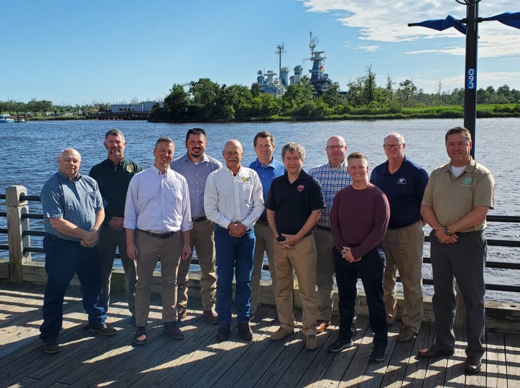 A group of individuals in business attire post on a boardwalk in front of a body of water, with a battleship in the backgroun