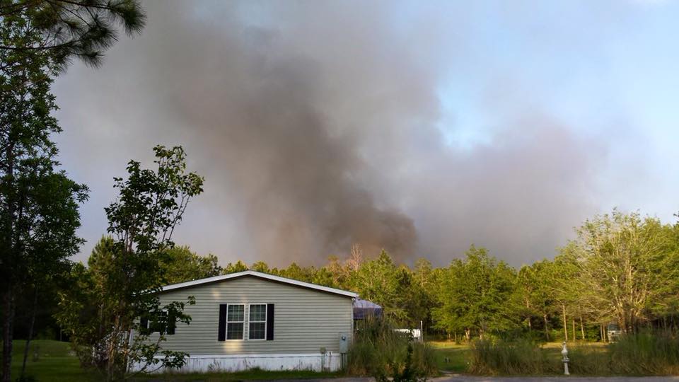 Mobile home in front of a wooded area that is on fire and producing smoke