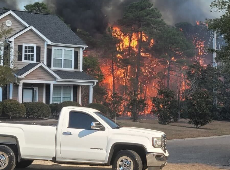 Wildfire raging behind a two story home, white pickup truck in the foreground