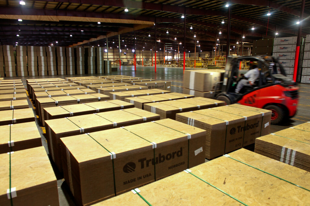 A high-ceilinged warehouse interior with rows of neatly stacked bundles of wood panels branded “Trubord – Made in USA” in the foreground, and a red forklift in motion loading panels on the right, with tall stacks of additional material and industrial lighting extending into the background.