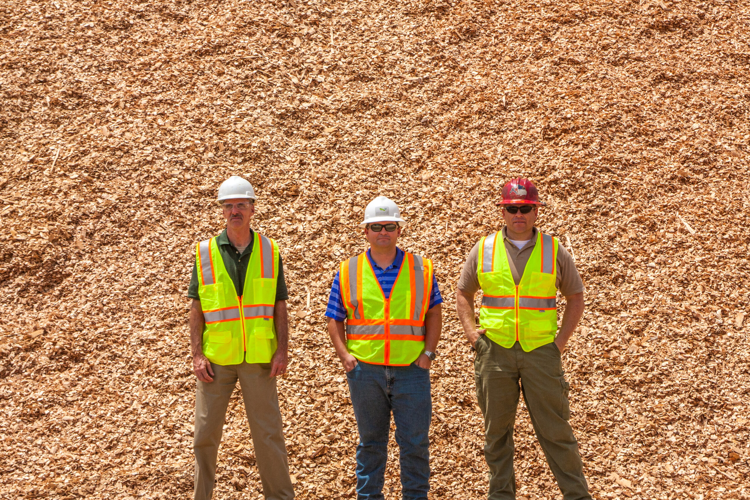 Three workers wearing hard hats and reflective safety vests stand in front of a large mound of wood chips.