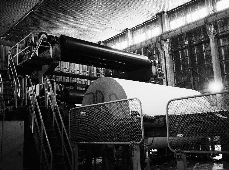 A large industrial paper mill machine in a high-ceilinged factory produces a massive roll of paper, with metal walkways, railings, and overhead equipment surrounding the machinery.