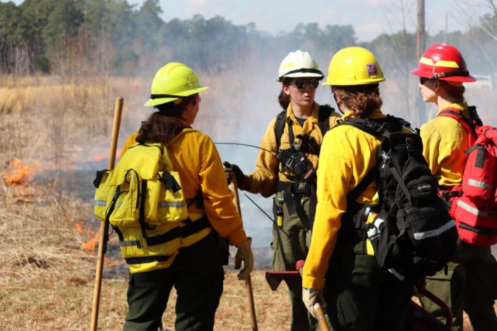 Group of female wildland firefighters converse while a prescribed fire burns gently in the background in a grass field. 