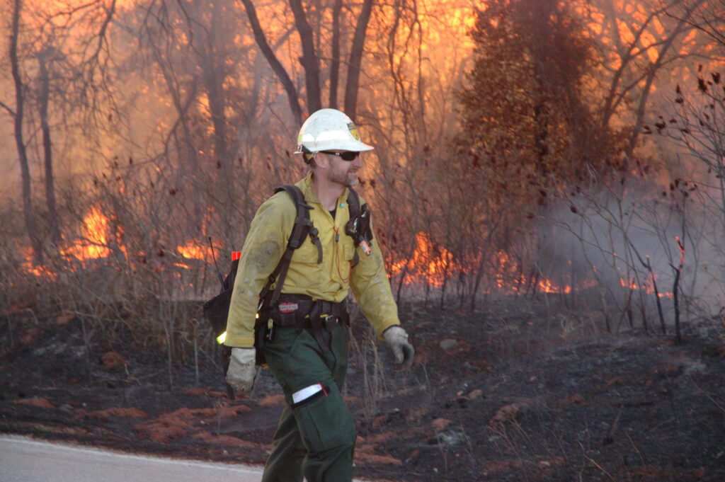 Wildland Firefighter walks alongside a fireline with burning debris in the background in an Oklahoma forest