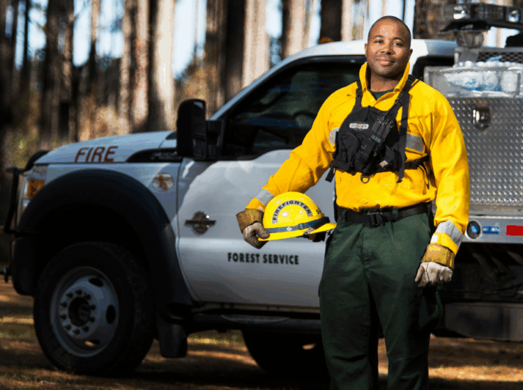 African American Wildland Firefighter poses in PPE, holding hard hat, standing in front of a brush truck and pine forest