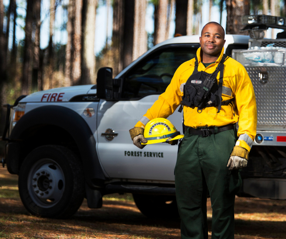 African American Wildland Firefighter poses in PPE, holding hard hat, standing in front of a brush truck and pine forest