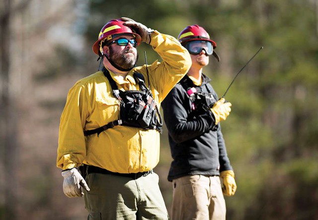 Two wildland firefighters outdoors, in PPE, looking off to the left. One is holding a radio with a long antenna.
