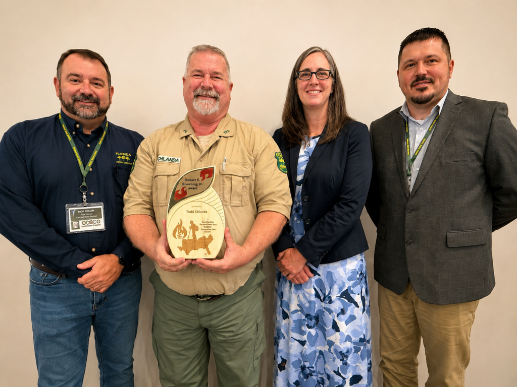 Four forestry professionals pose for a recognition photo against a light-colored wall. At center, Todd Chlanda holds the Robert E. Browning Jr. Award, a decorative plaque recognizing excellence in wildfire prevention. He is flanked by colleagues wearing conference badges and business attire. The group is smiling and facing the camera.