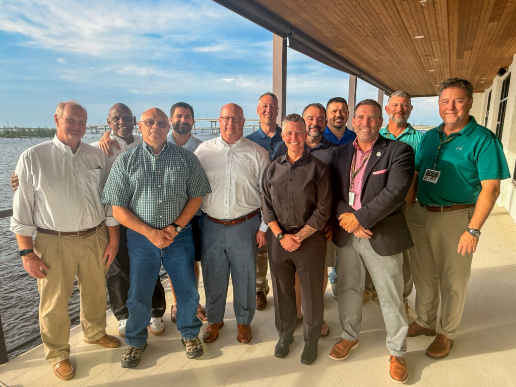 A group of 11 attendees stands together on a covered waterfront terrace during golden hour, posing for a photo with a bay and bridge visible in the background. The group is dressed in a mix of business casual and casual attire, with sunlight illuminating their faces. The setting overlooks calm water and coastal scenery, creating a relaxed atmosphere during an evening networking event.